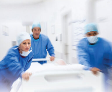 Photo of three healthcare workers wearing scrubs and pushing a gurney in an ER